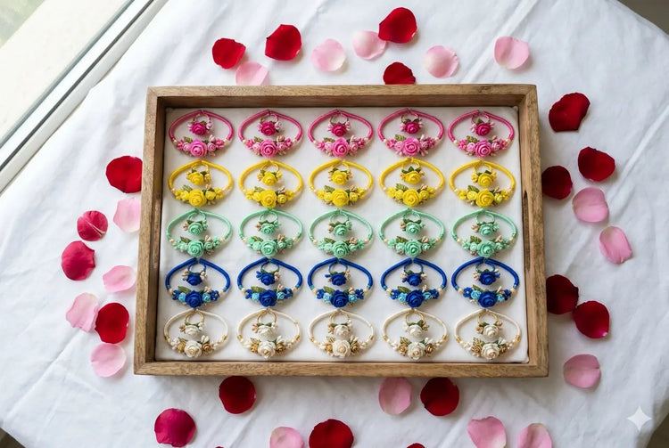 Colourful floral ring and bracelet combos in pink, yellow, green, blue and pearl white arranged neatly in a wooden tray on white cloth with rose petals scattered around for wedding return gift packaging
